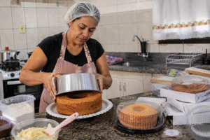Mulher preparando bolo caseiro para vender em cozinha doméstica, com bolos embalados sobre a bancada, representando renda extra e empreendedorismo na confeitaria.
